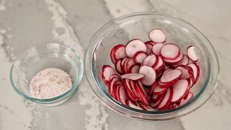 Sliced radishes in a glass bowl sitting next to a small bowl of unrefined salt